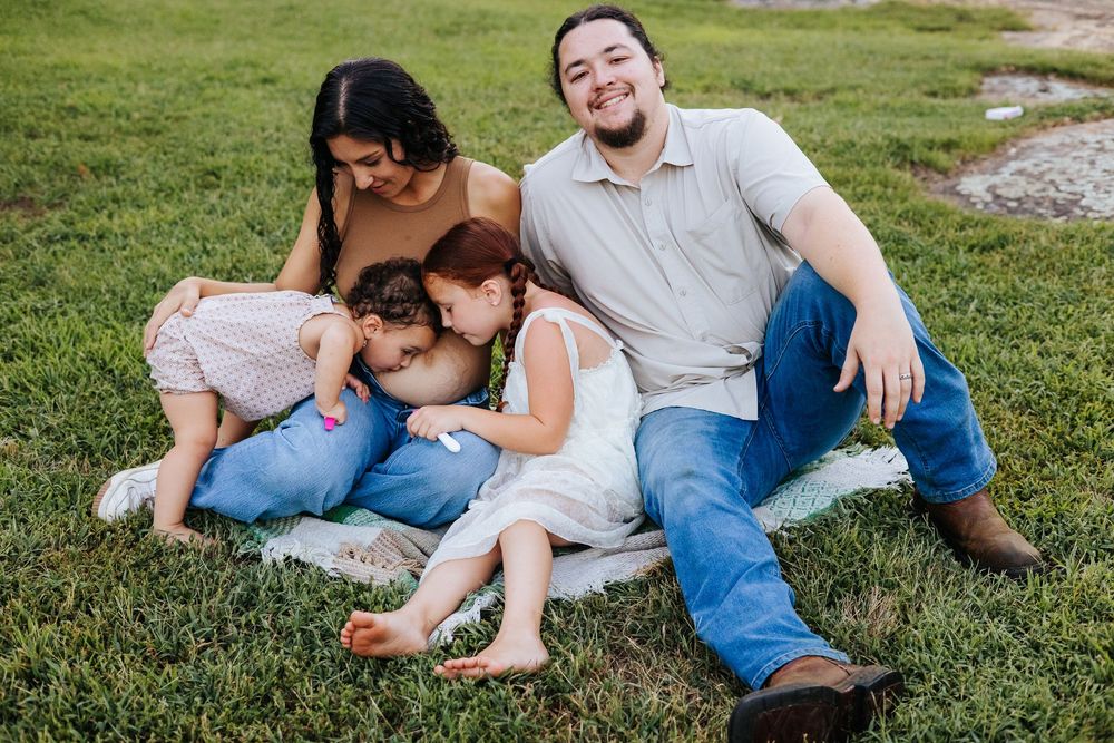 Happy family of four sitting on grass, parents smiling as two young children cuddle together on a sunny day.