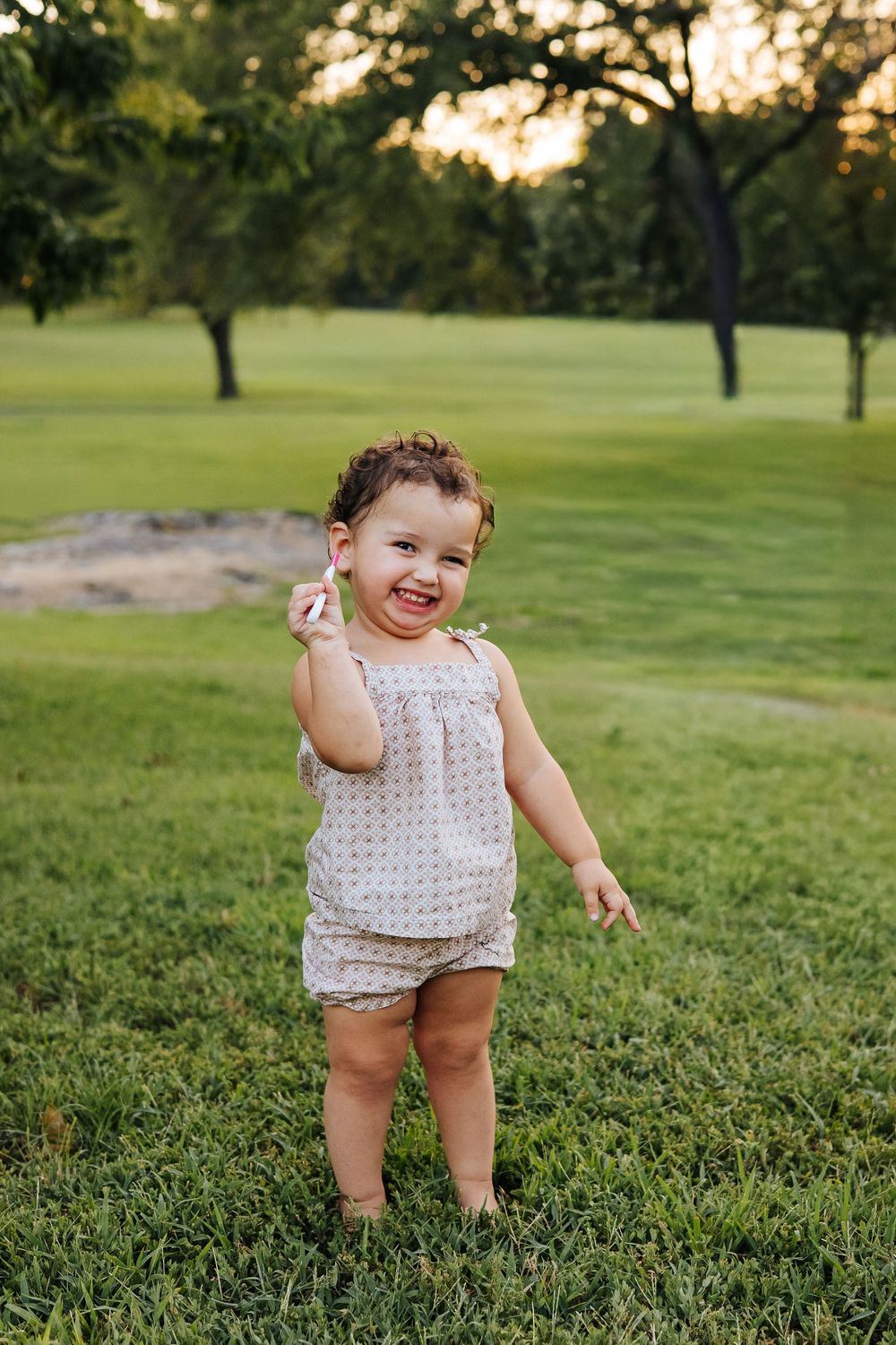 Happy toddler girl smiling and standing barefoot on green grass in a park at sunset.