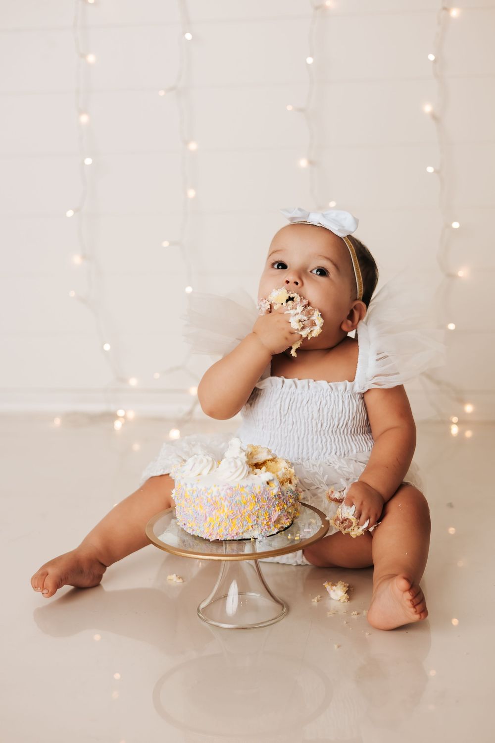Cake smash photo session with twinkling lights and white cake on stand.