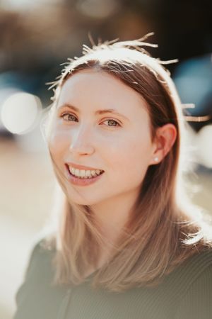 Portrait photo of someone with straight hair smiling warmly in natural sunlight with soft bokeh in background.