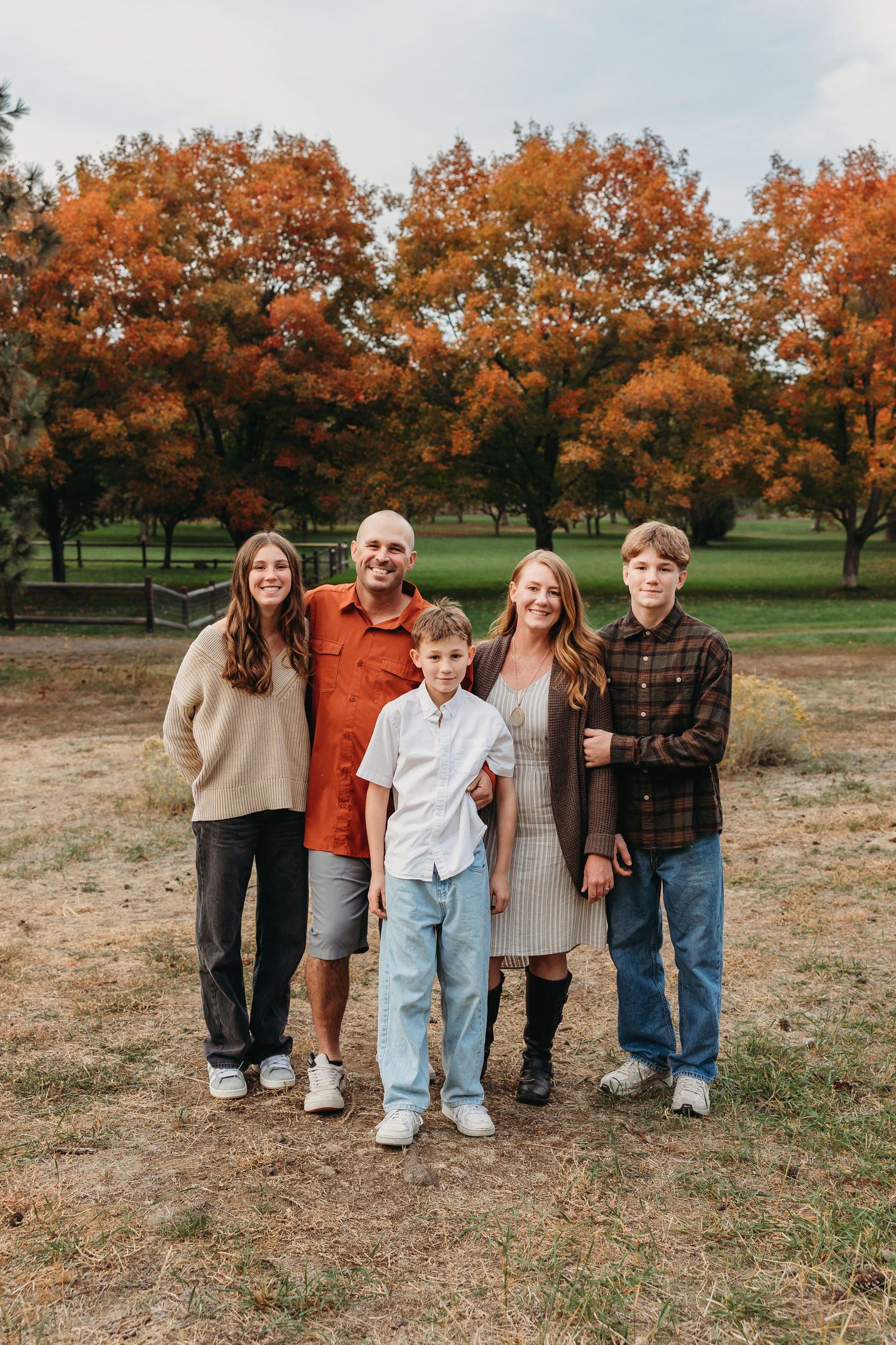 Family group poses together on grass field with autumn orange trees in background during fall photo session.