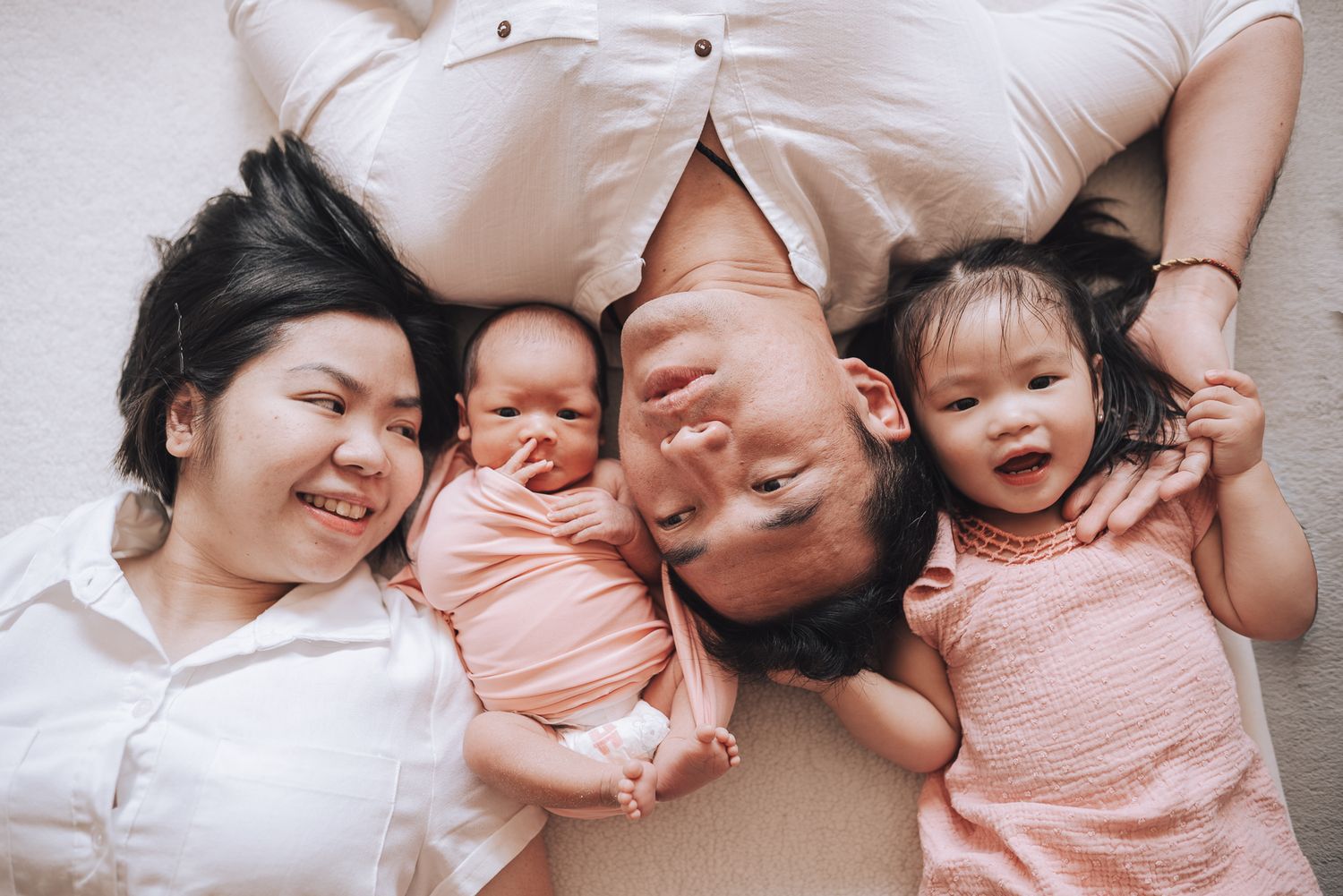 A happy family of four lying together on white bedding while smiling and laughing taken during an in-home Melbourne newborn photo session.