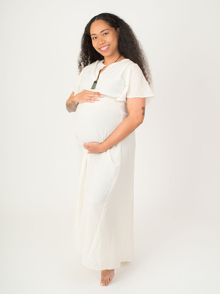 Photo of a pregnant woman with curly hair wearing a flowing white dress standing barefoot against a plain background by Abigail's Studio