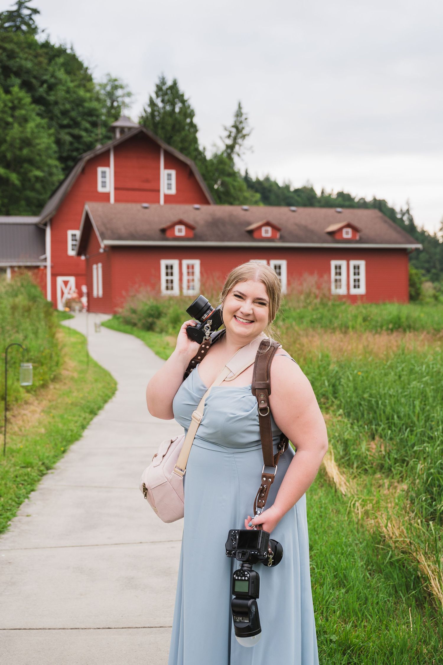 A photographer in a blue dress stands on a concrete path with red barns and green fields in the background.