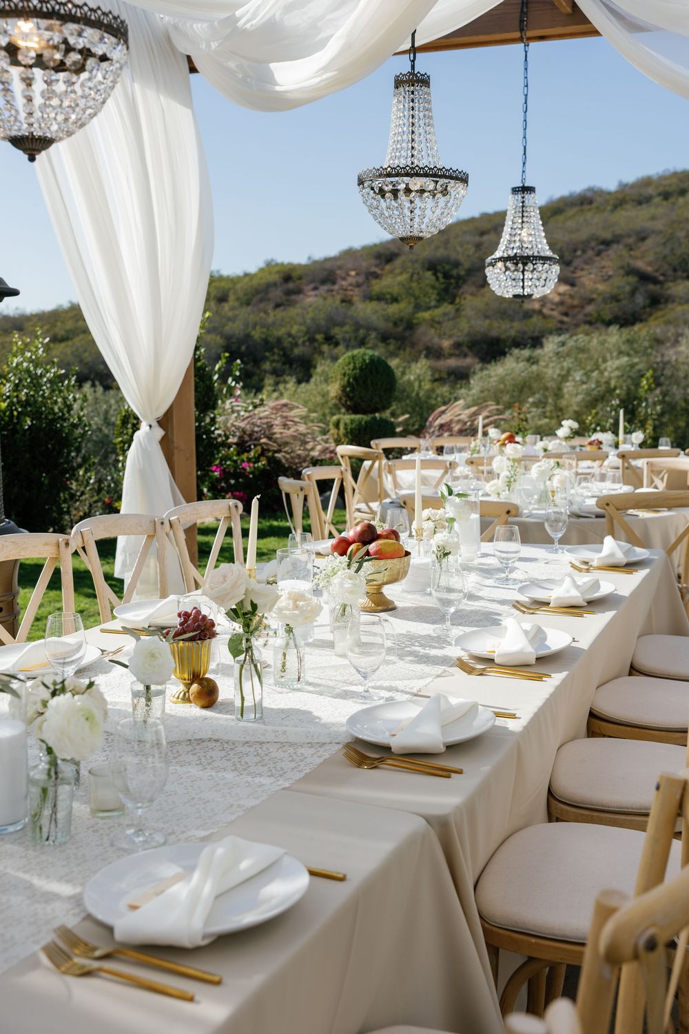 Elegant white reception tables with crystal chandeliers and draped fabric in an outdoor tent setting.