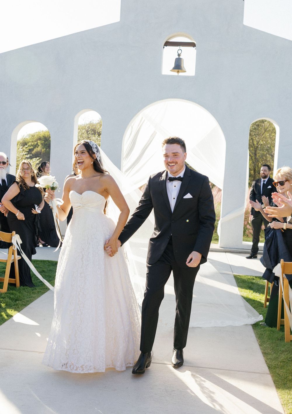 Newlyweds joyfully exit their wedding ceremony under a white archway while guests look on.