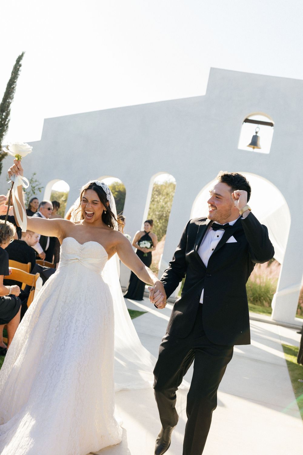 A joyful wedding couple exits their ceremony under white arches in bright sunlight.