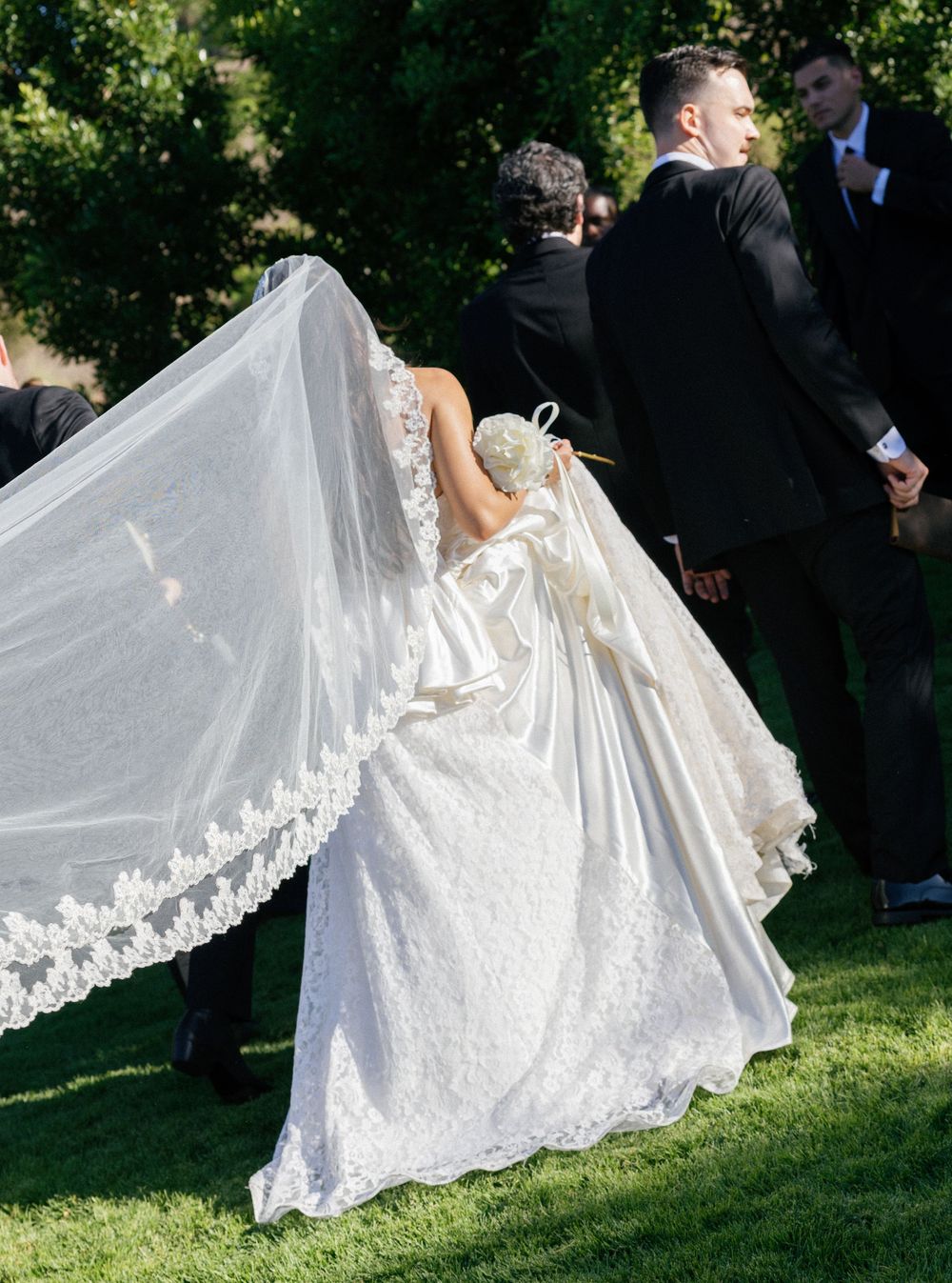Elegant wedding dress with flowing lace train spreads across green lawn during outdoor celebration.