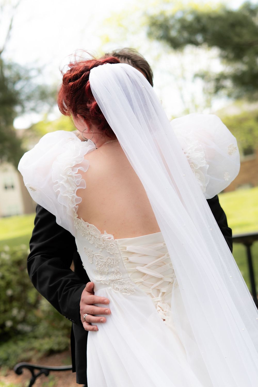 Bride and groom sharing tender embrace outdoors, showcasing elegant lace wedding dress with long veil.