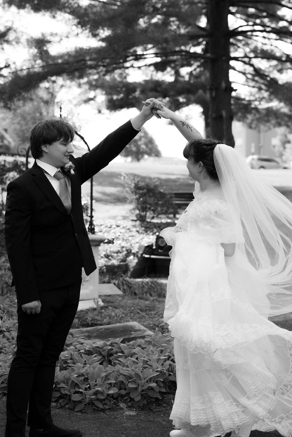 Black and white photo of groom spinning bride in wedding dress and veil in outdoor garden setting.