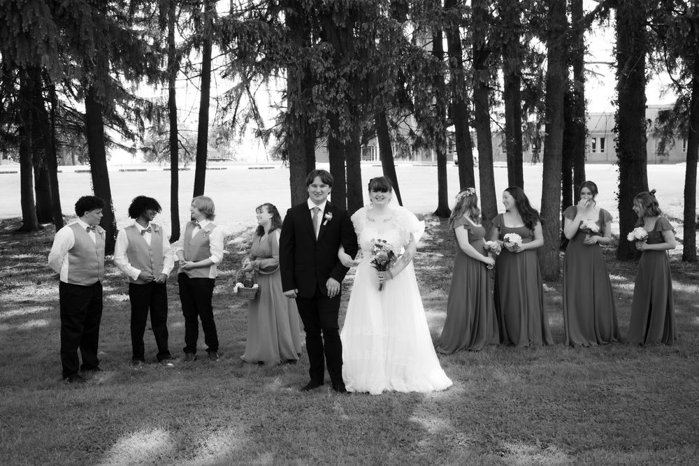 Black and white wedding party portrait with bride, groom, and bridesmaids in long dresses among pine trees.