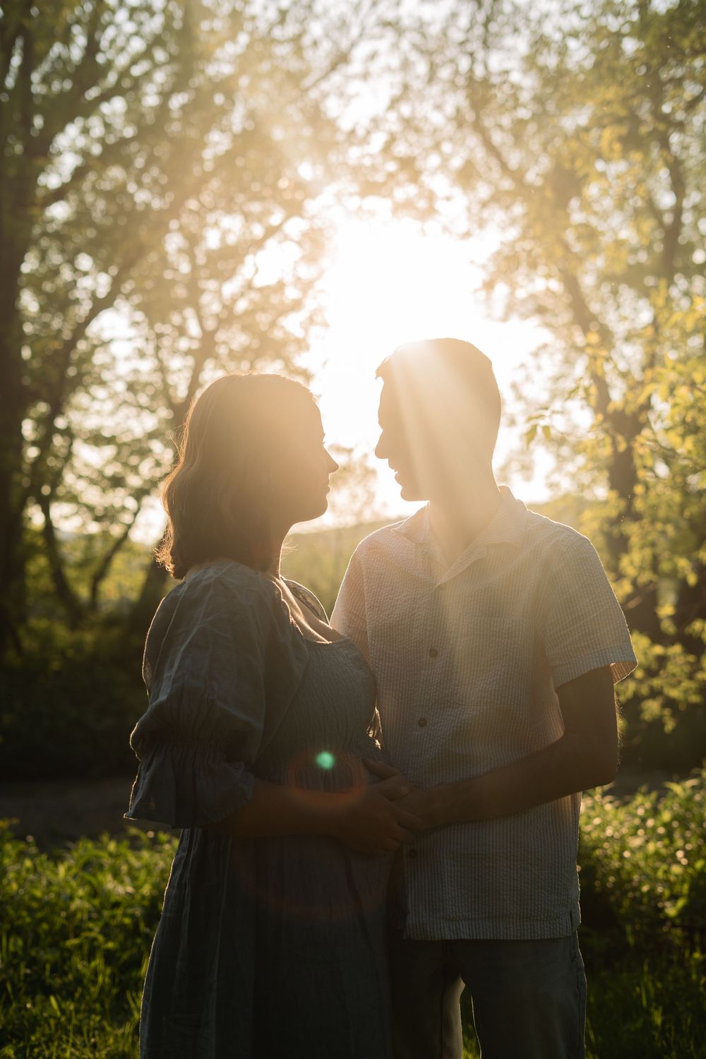 A romantic couple silhouetted against golden sunlight embrace face-to-face in a sunlit green forest setting.