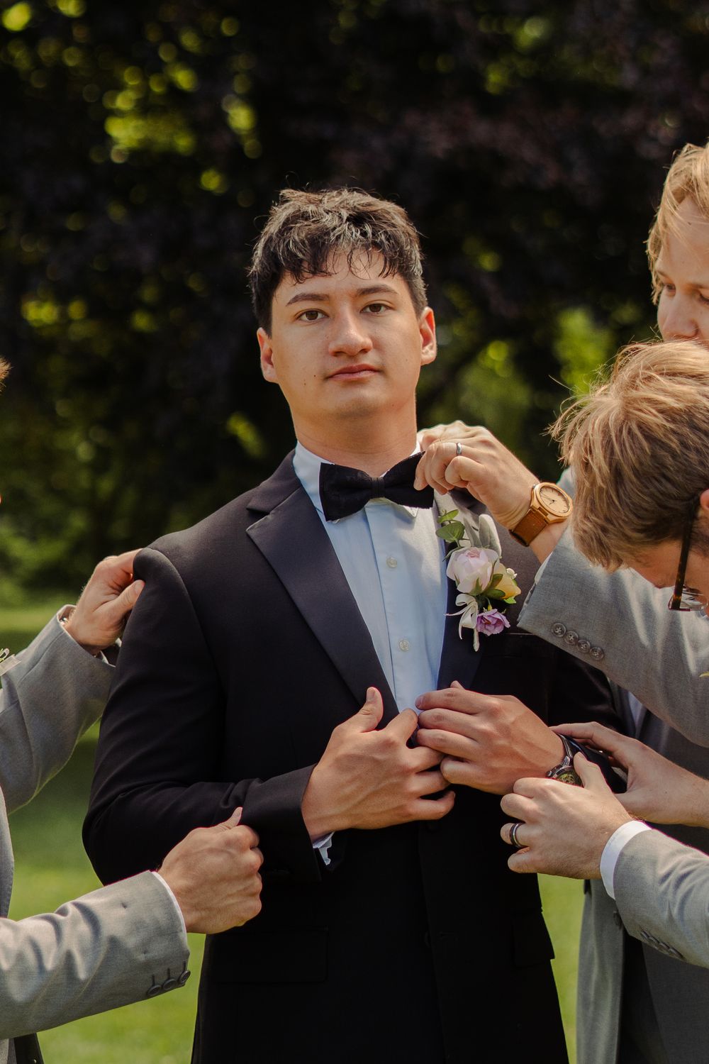 Groomsmen help groom adjust his tuxedo and boutonniere outdoors before wedding ceremony.