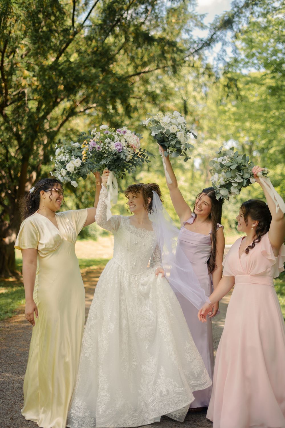 Bride and bridesmaids in pastel gowns raise bouquets joyfully under tree-lined path.