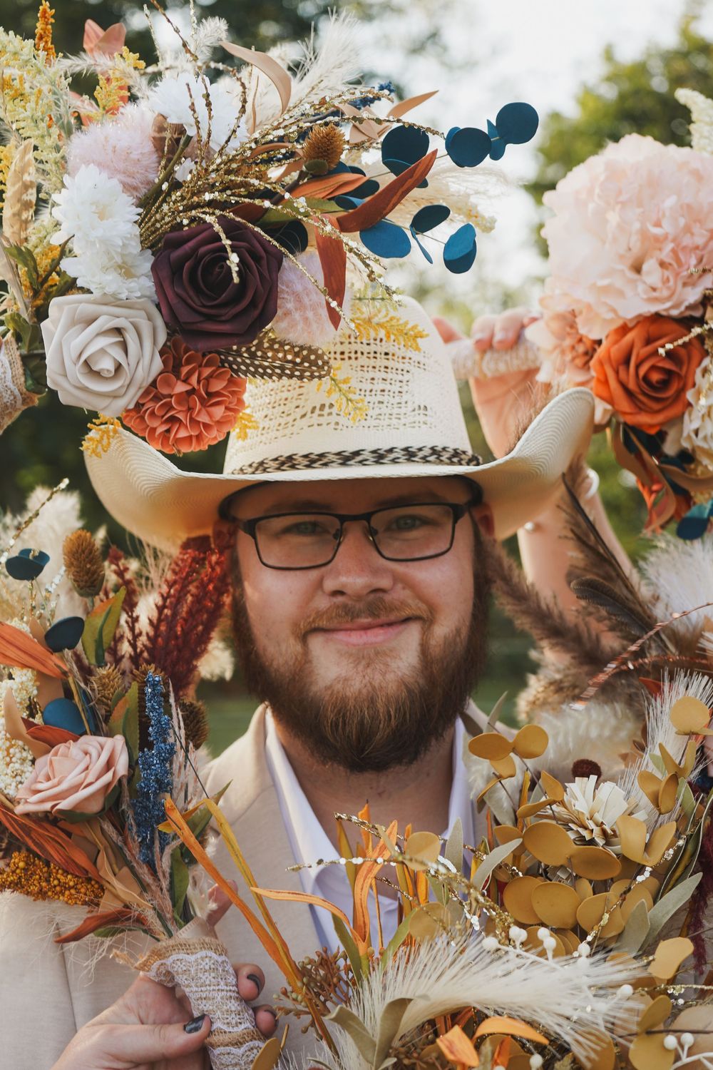 Smiling bearded groom in cowboy hat surrounded by colorful boho floral arrangements at outdoor wedding.