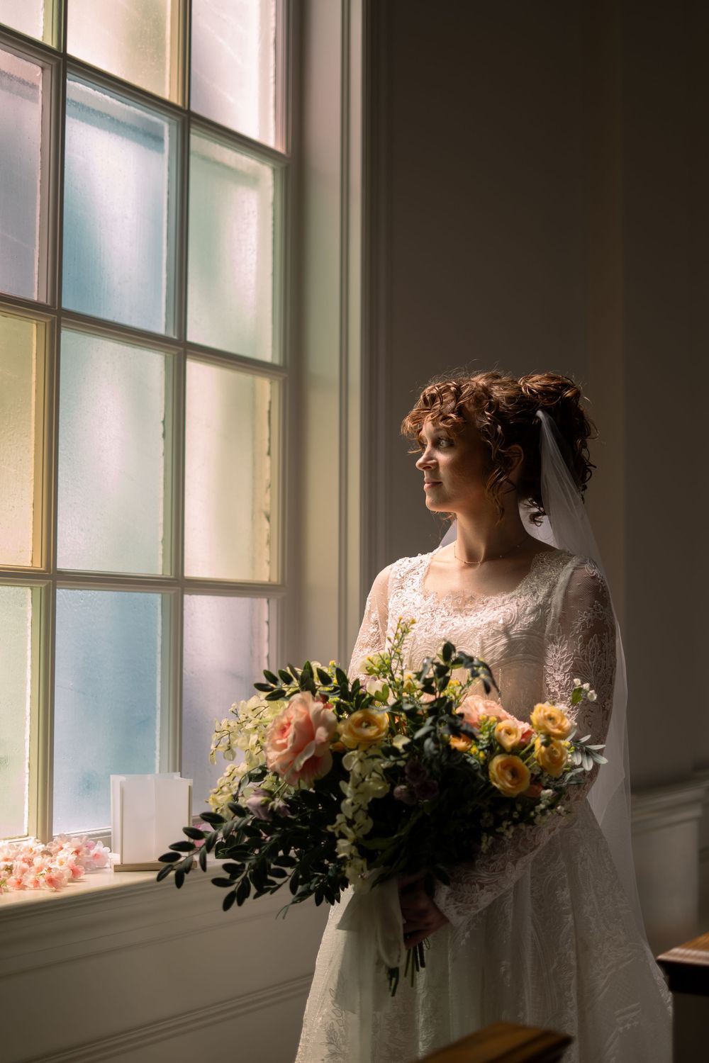 Bride in lace gown holds floral bouquet by stained glass window in soft natural light.