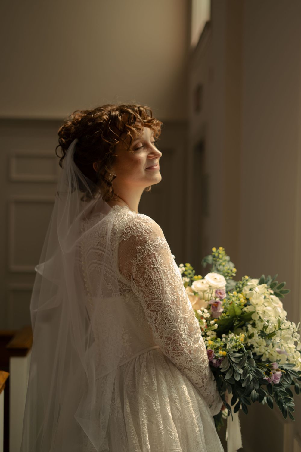 Elegant bride in lace gown with veil holds bouquet in softly lit indoor setting.