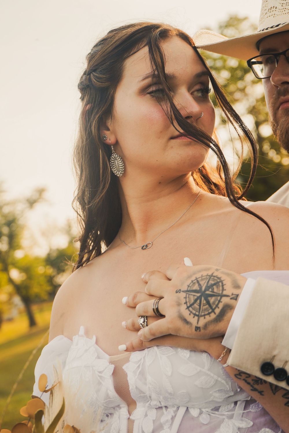 Bride in white off-shoulder dress with compass tattoo hand resting on chest at golden hour outdoor wedding.