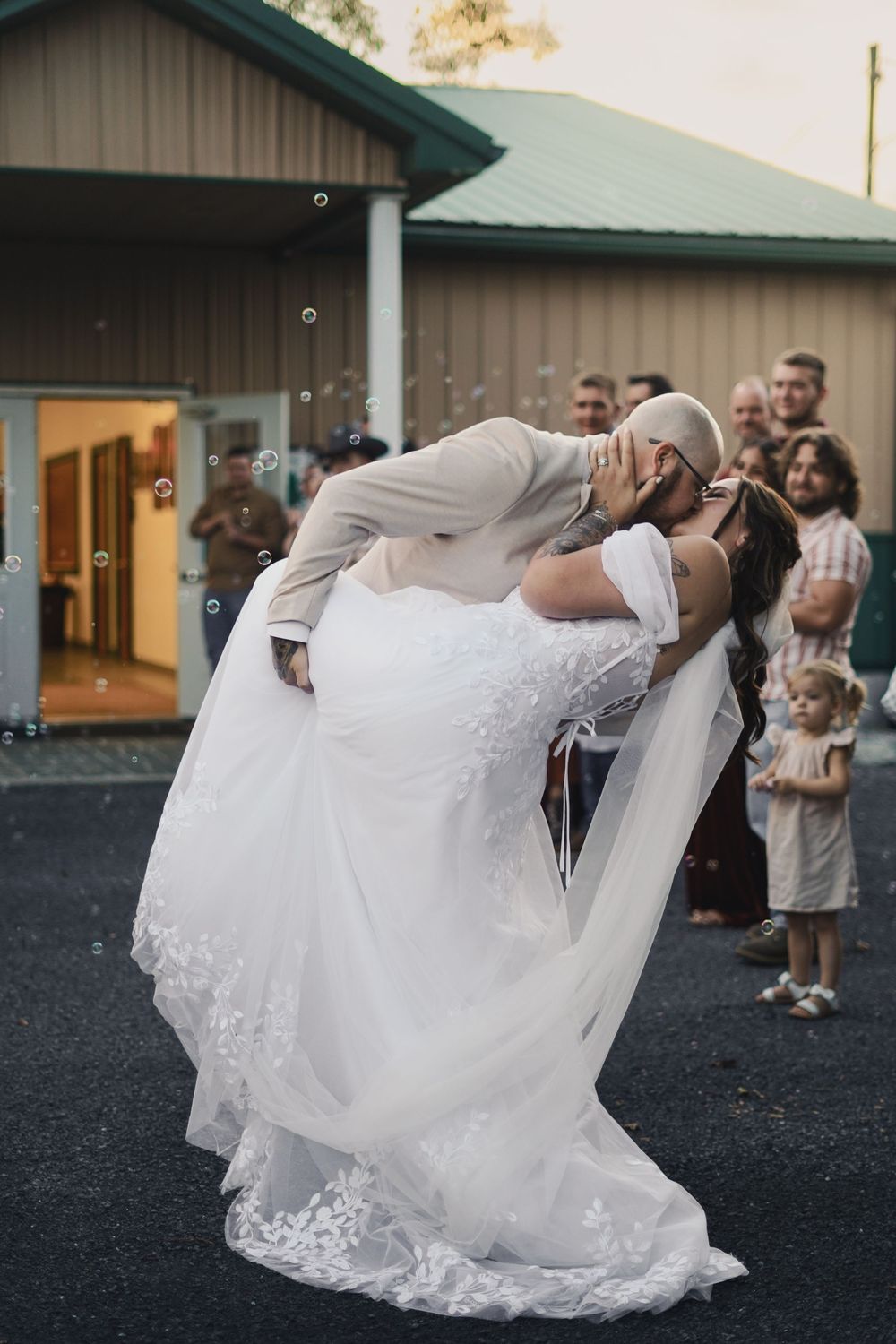 Groom lifts and dips bride in white lace wedding gown for romantic kiss outside venue with bubbles floating.