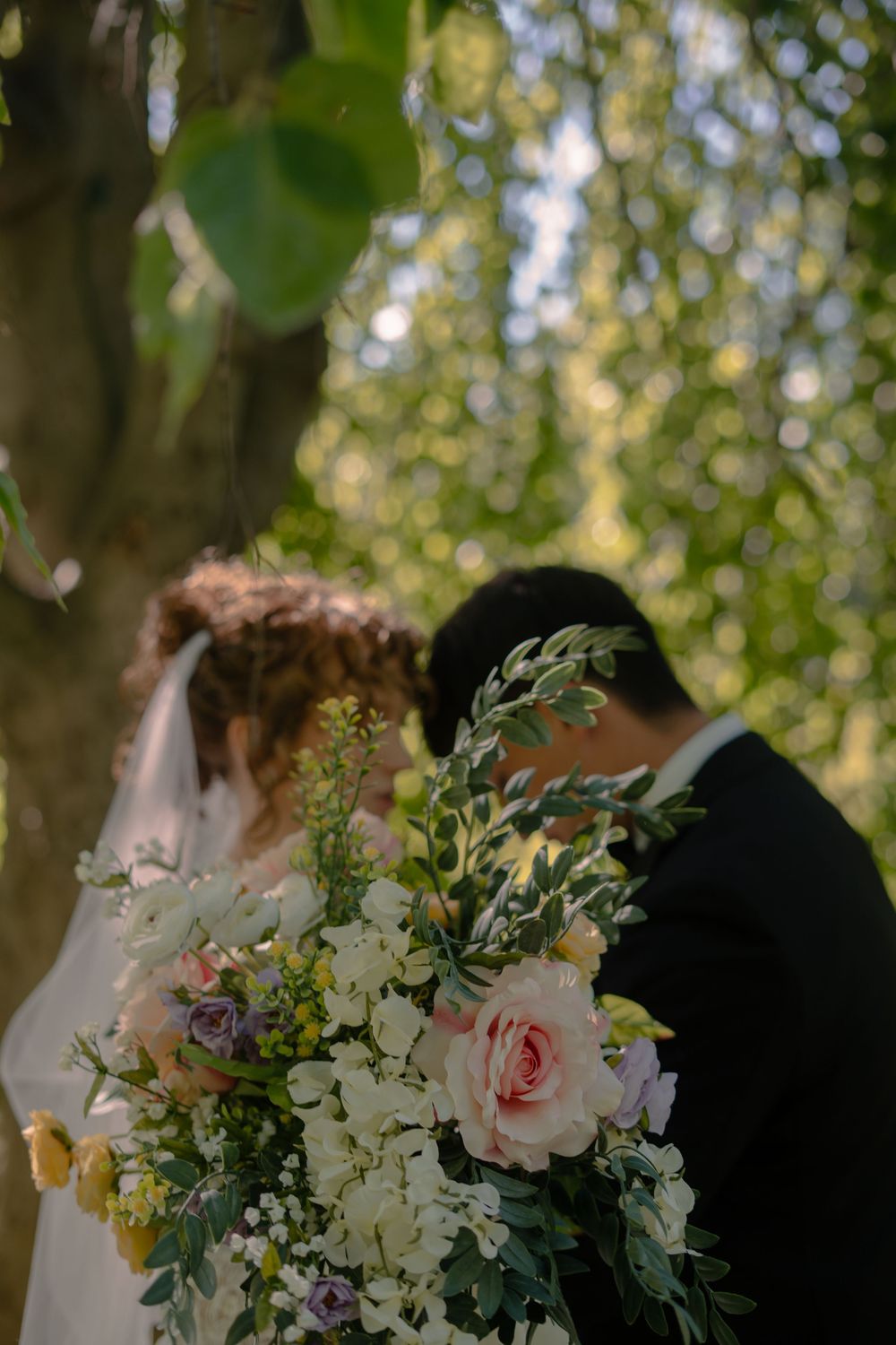Bride and groom hidden behind lush wedding bouquet of roses and greenery outdoors.