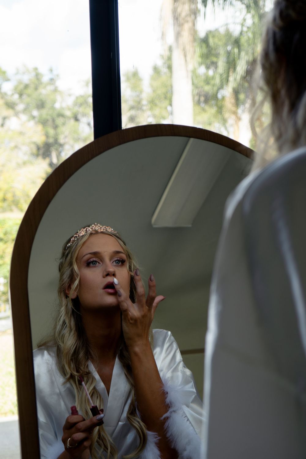 Blonde bride in white robe applying lipstick while looking at reflection in round mirror during wedding prep.