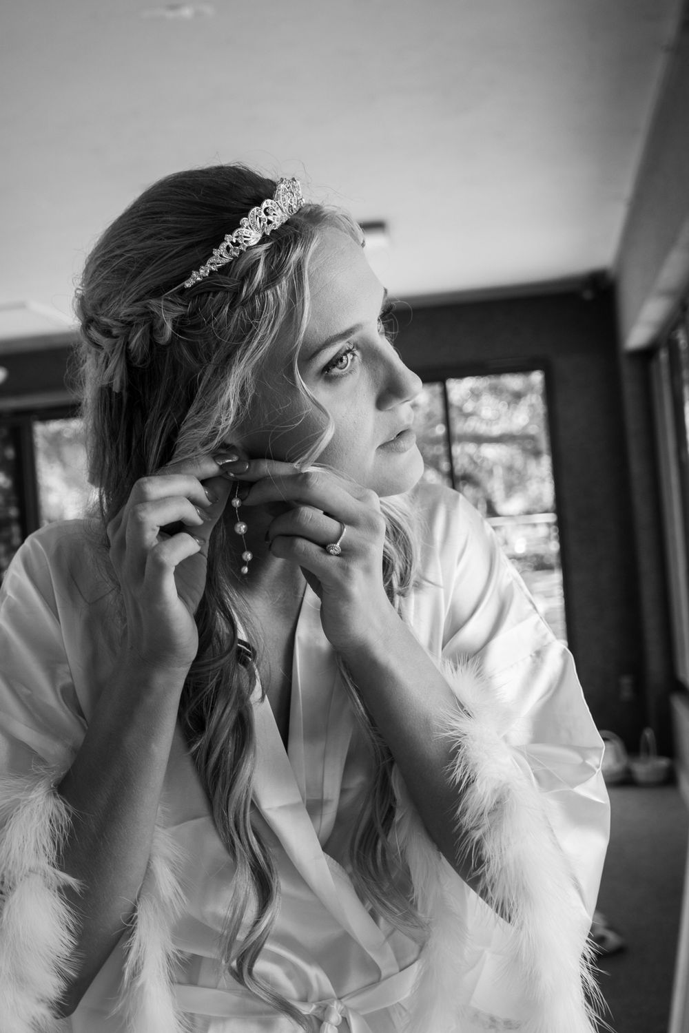 Black and white portrait of bride in white robe wearing tiara and pearl earrings during wedding preparations.