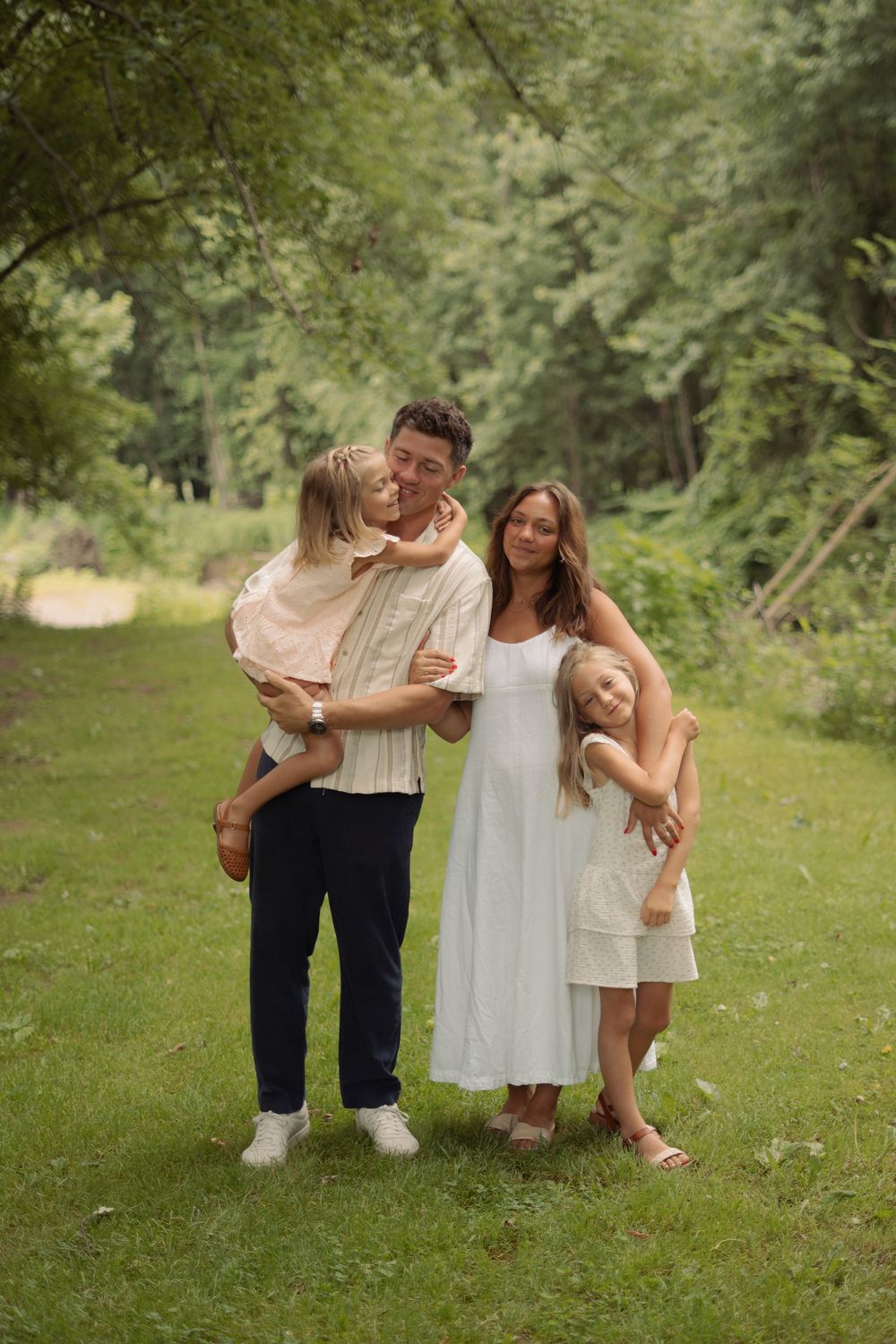 Happy family of four embraces on a green forest path during outdoor photo session.