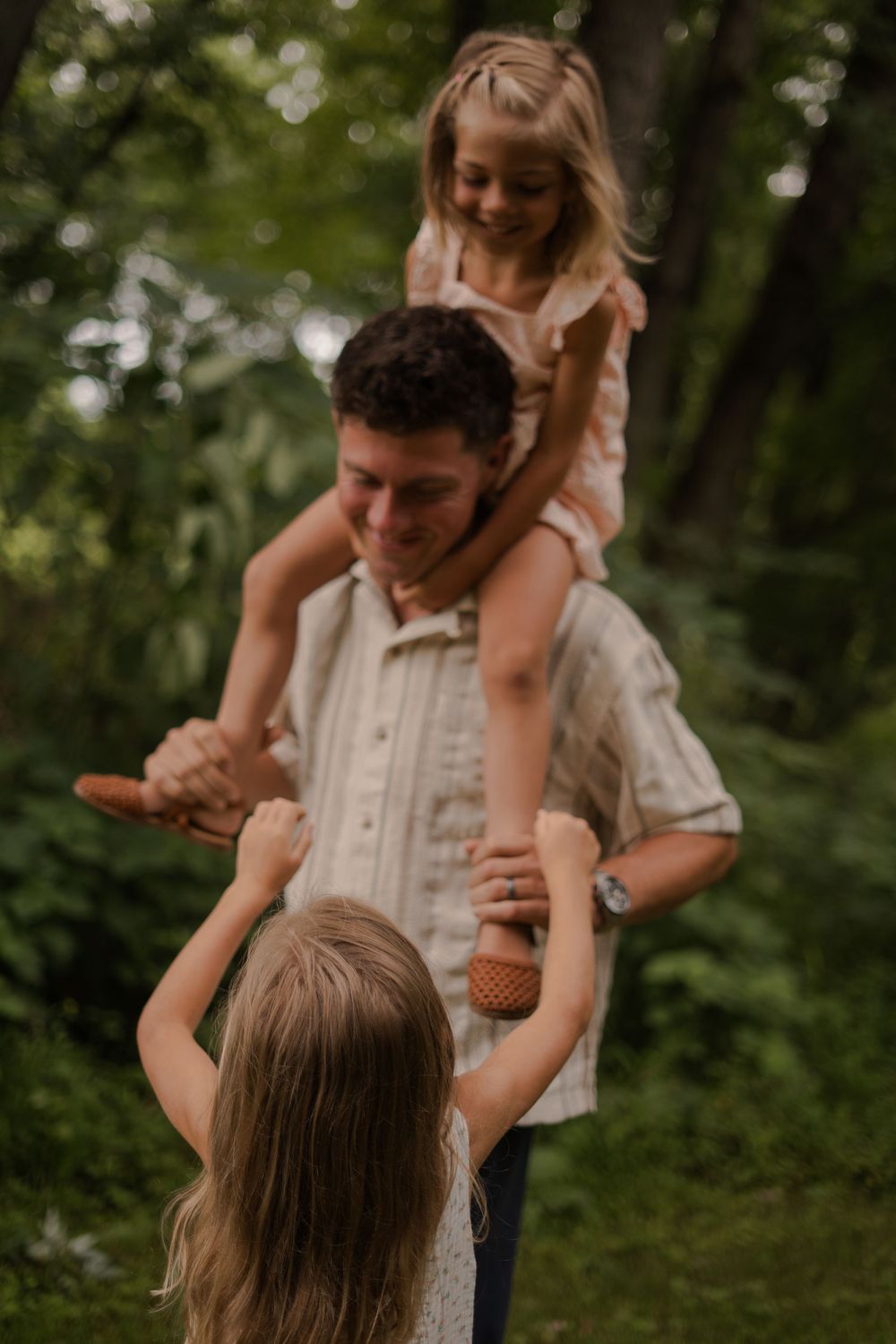 Father with daughter on shoulders plays with two girls in a lush green forest setting.