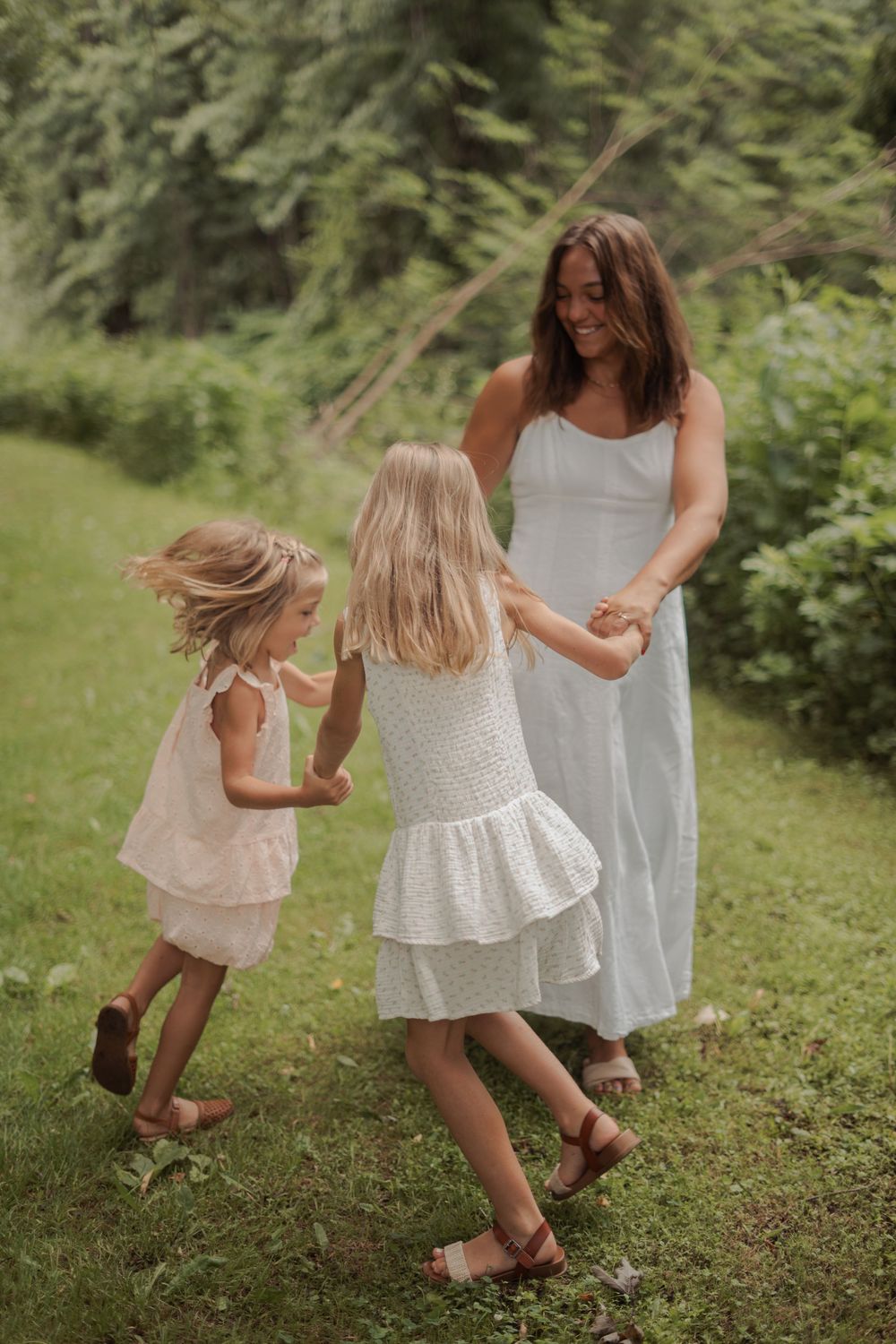 A mother in a white dress watches her two young daughters in white dresses hold hands and dance in a lush green outdoor setting.