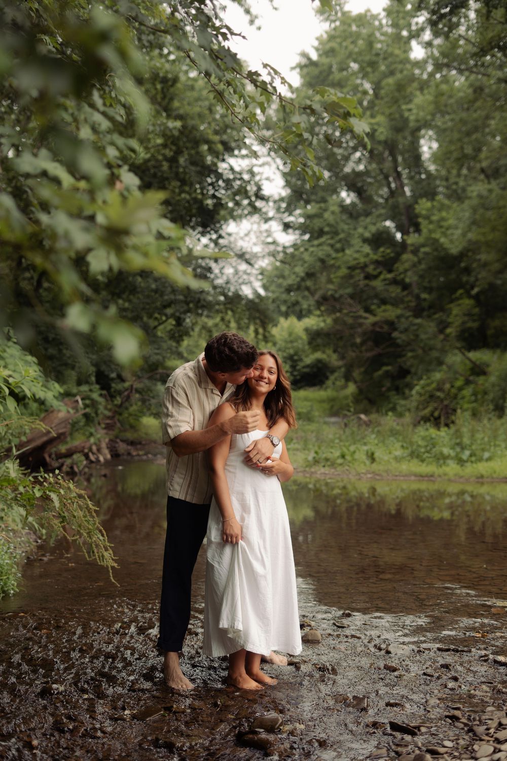 A couple embraces and laughs together while standing barefoot in a shallow creek surrounded by lush green trees.