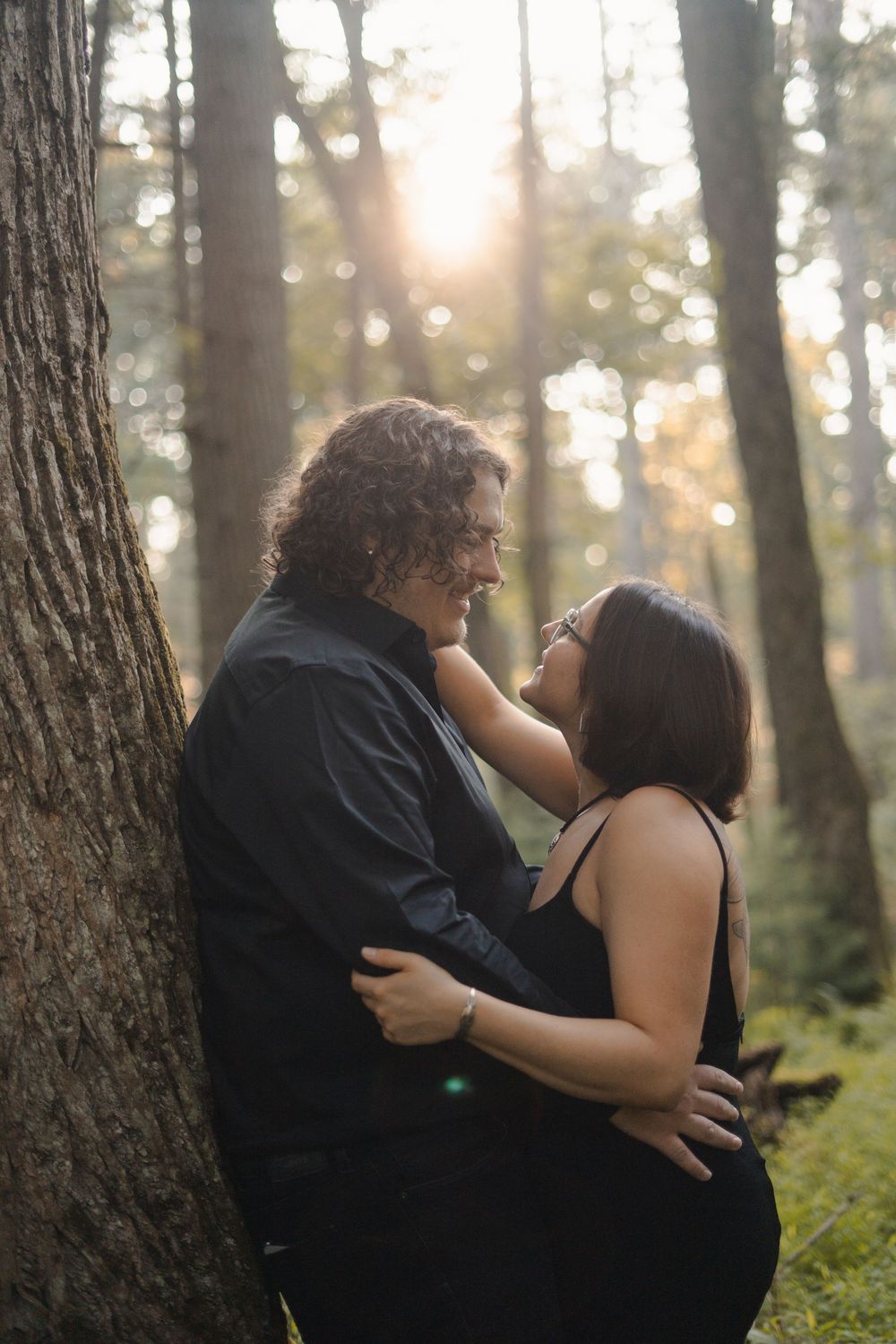Couple embracing romantically against a tree in a sunlit forest, sharing a tender moment outdoors.