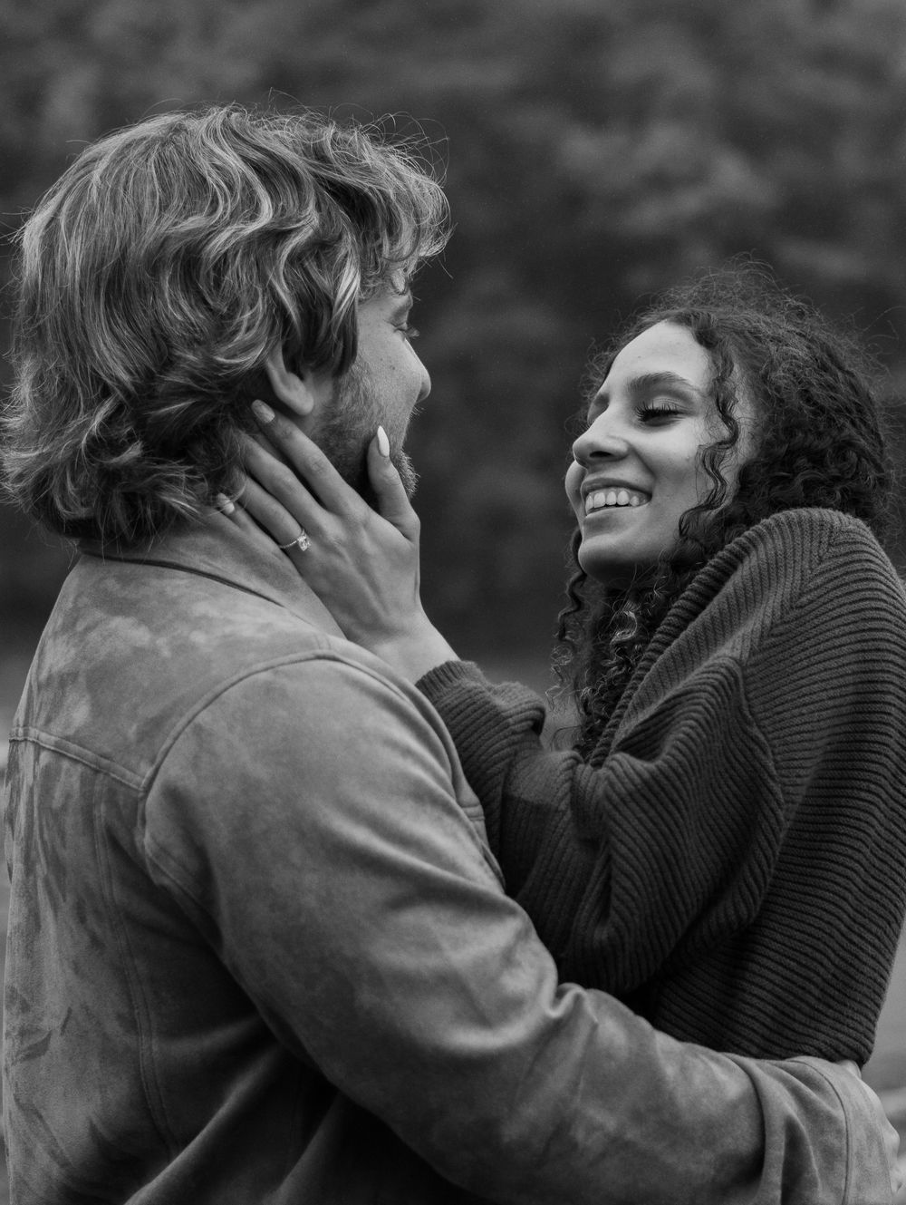 A smiling woman tenderly cups a mans face in a black and white outdoor couples portrait.