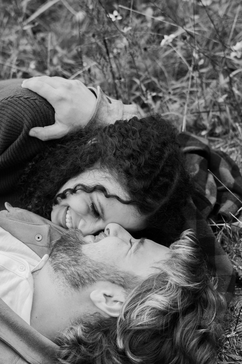 Black and white romantic couple lying in grass field, smiling and gazing at each other surrounded by wildflowers.