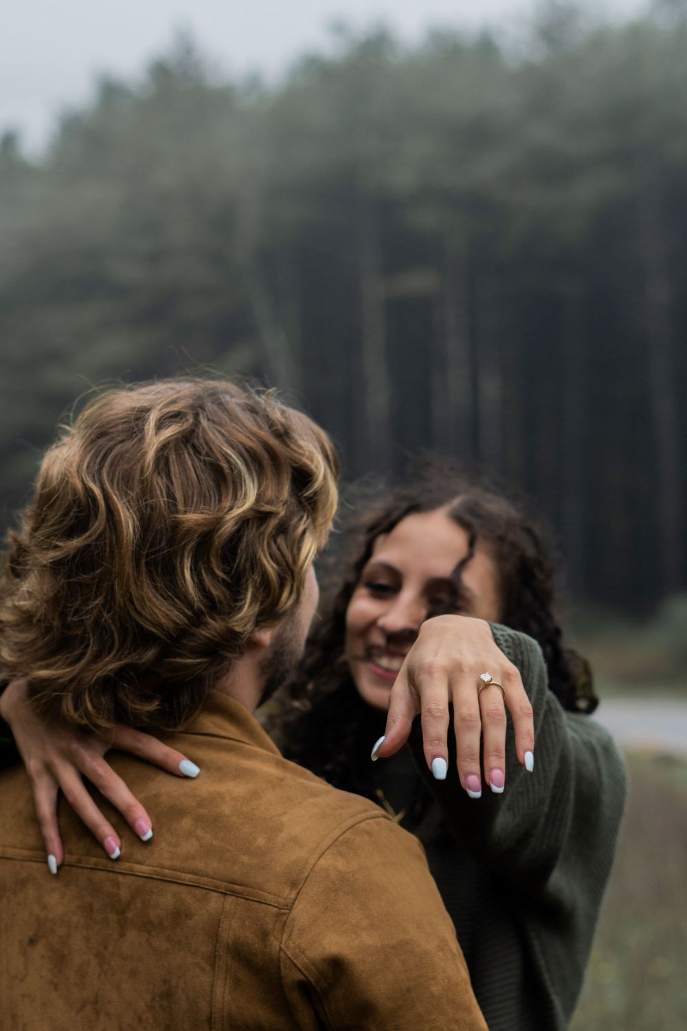 Woman smiling and showing off engagement ring while embracing partner outdoors near foggy pine forest.
