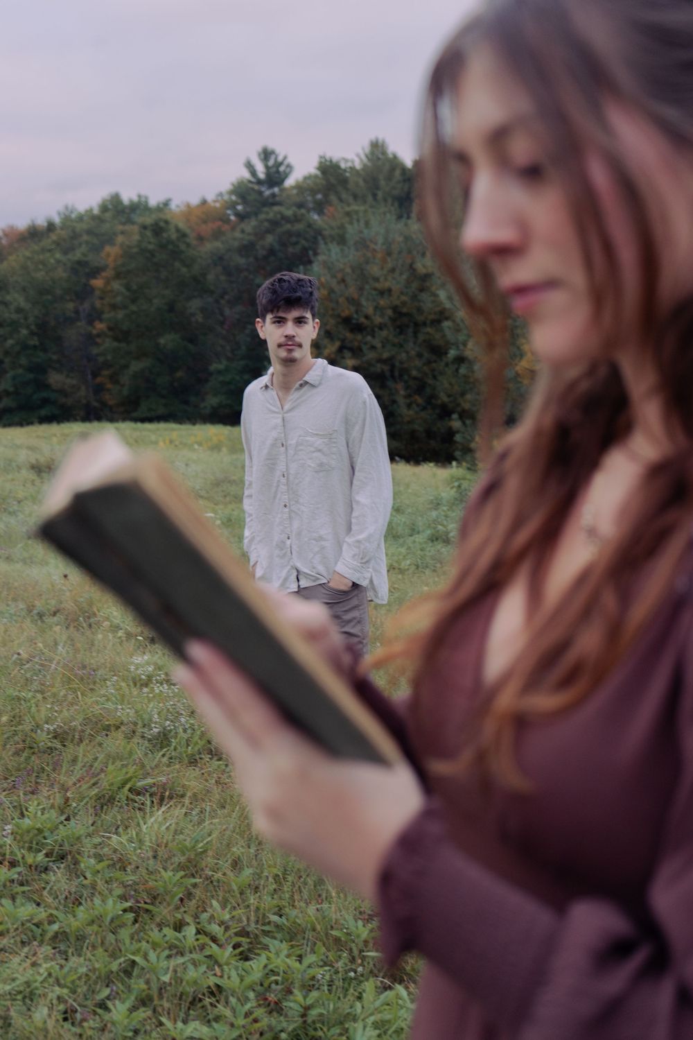 A woman in a maroon top reads an old book in a grassy field while a young man in a linen shirt stands blurred behind her.