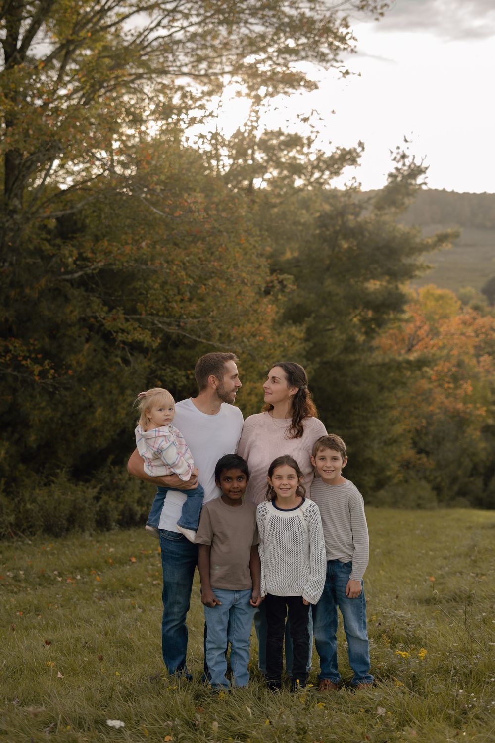 Family of six poses together in a grassy field, parents holding a toddler with three children in front, autumn trees behind them.