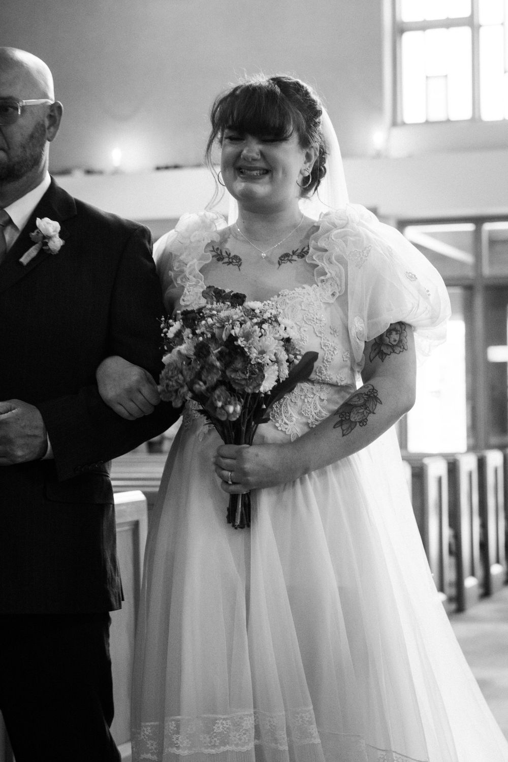 Black and white photo of smiling bride walking down church aisle holding floral bouquet in lace wedding gown.
