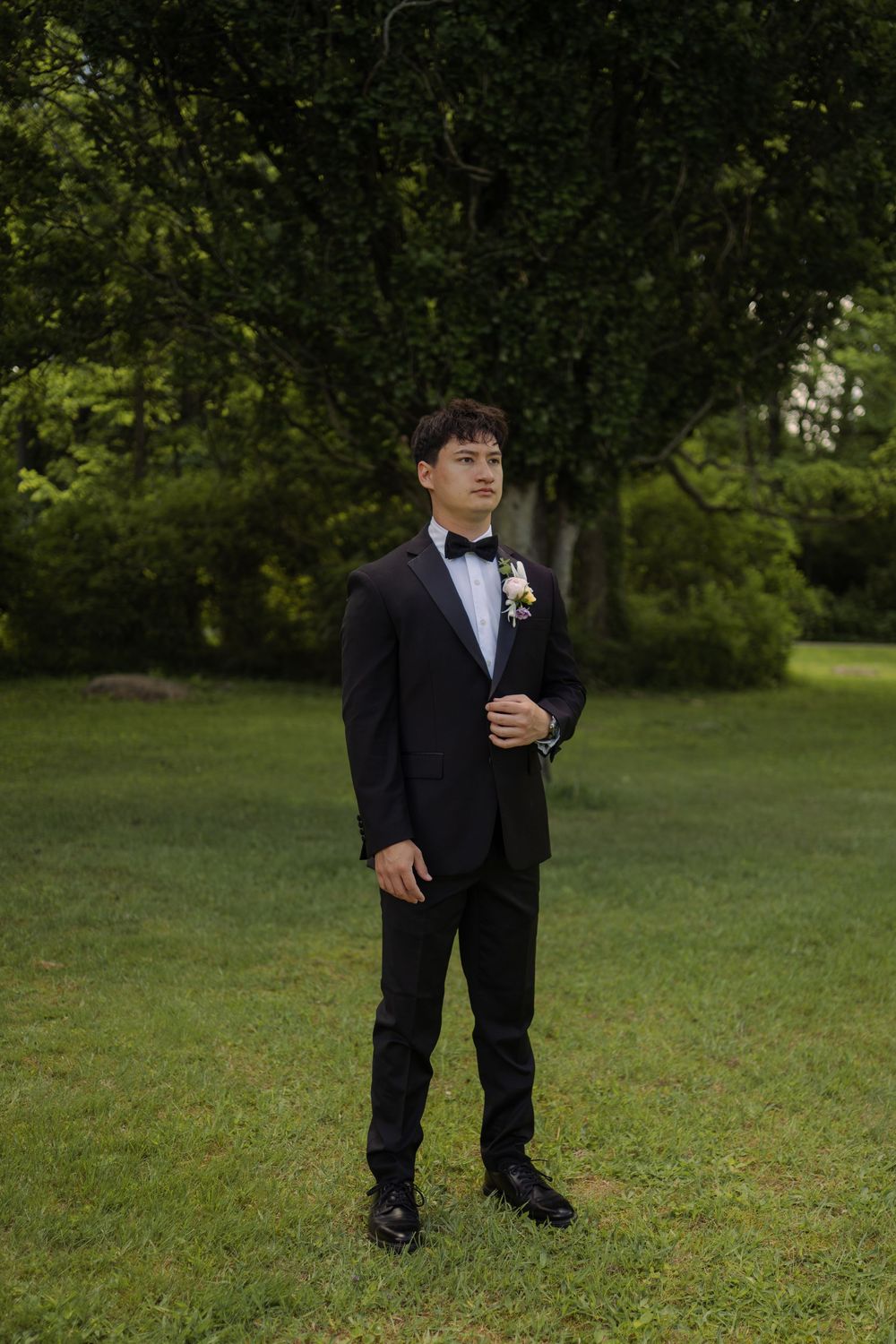 Groom in black tuxedo with boutonniere stands on green lawn surrounded by trees.