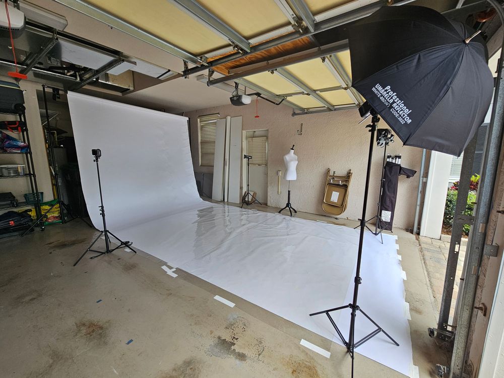 Photography studio setup showing white backdrop, lighting equipment, and stands arranged in a garage space.