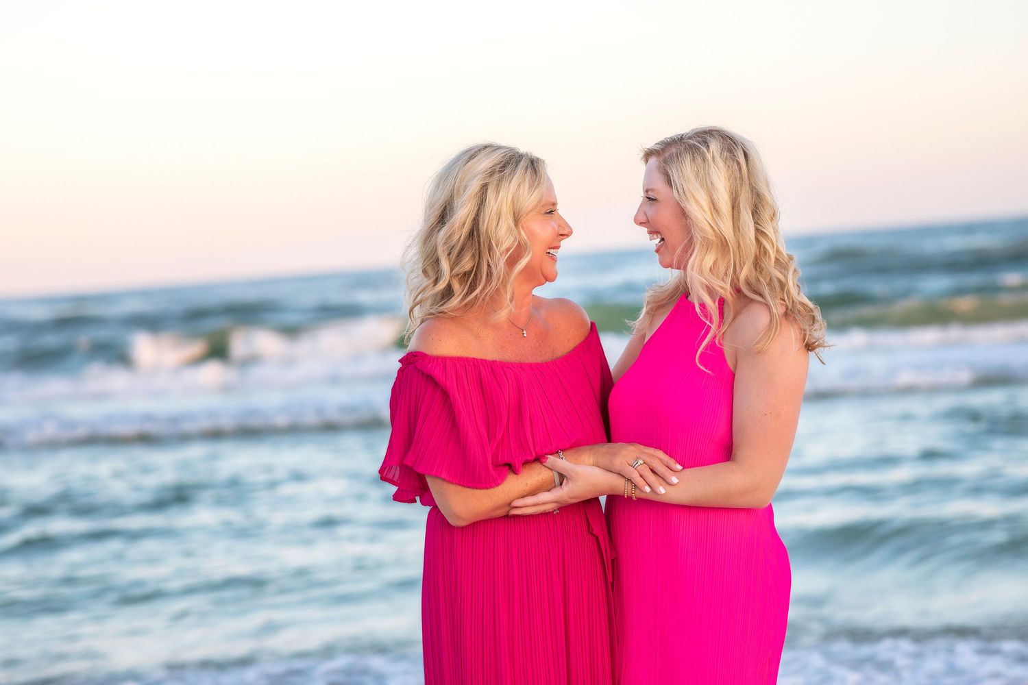 Two people in vibrant pink dresses embrace while walking along a beach at sunset.