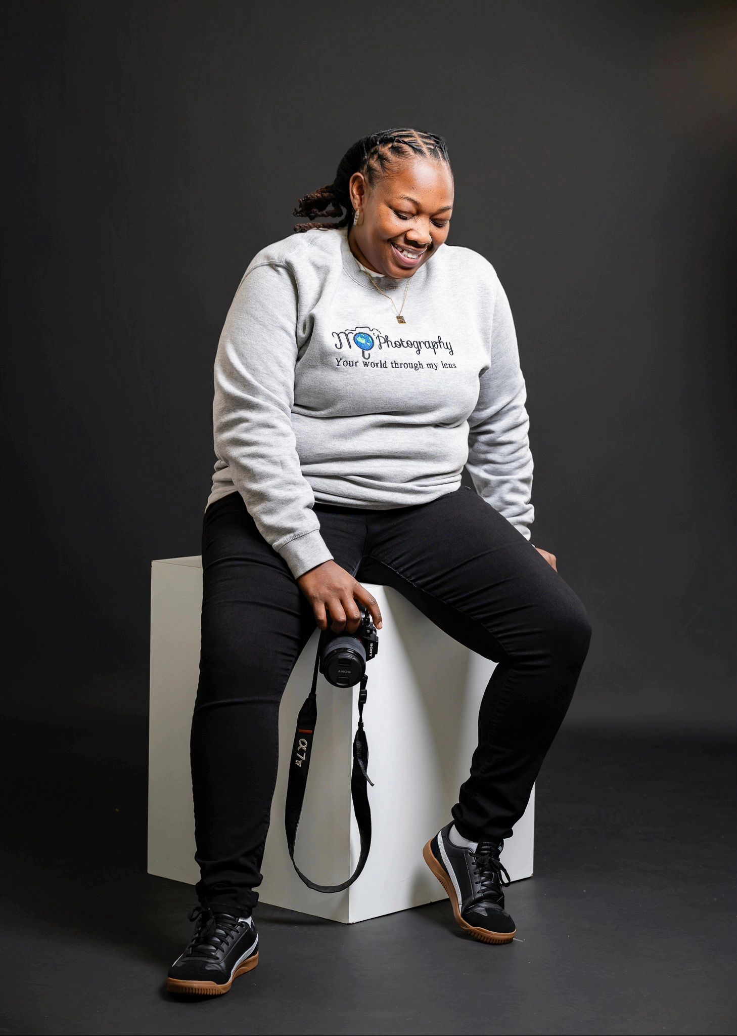 Person wearing grey sweatshirt and black pants sitting on white cube against dark backdrop.