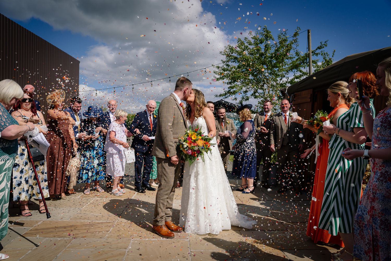 Wedding guests throw confetti at a joyful celebration on a sunny day with blue skies overhead.