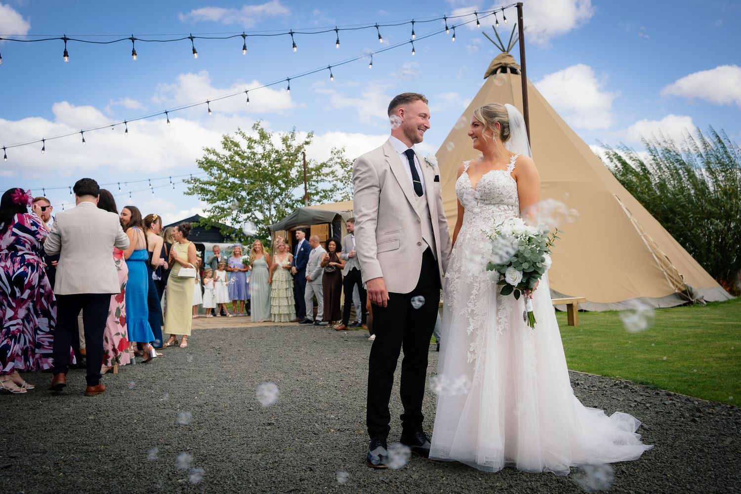 A couple celebrates their outdoor wedding reception by a tipi tent with string lights and guests gathered on a summer day.