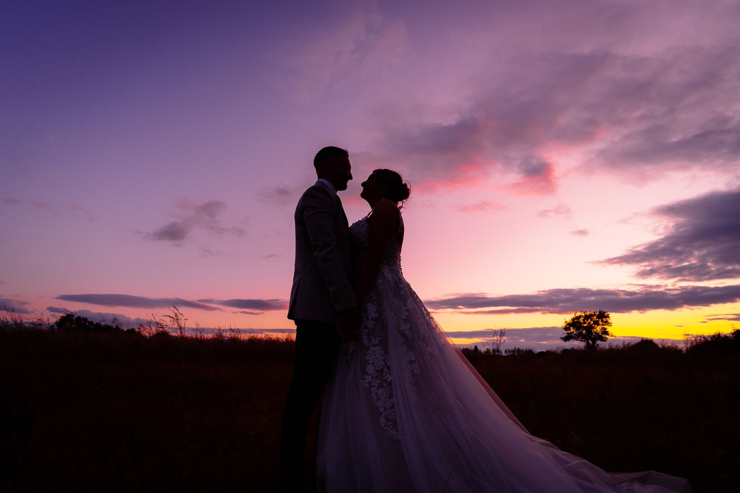 Silhouetted couple embraces against a vibrant purple and orange sunset sky with clouds in the background.