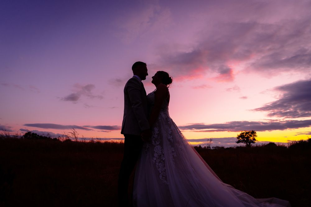 Silhouetted couple embraces against a vibrant purple and orange sunset sky with clouds in the background.