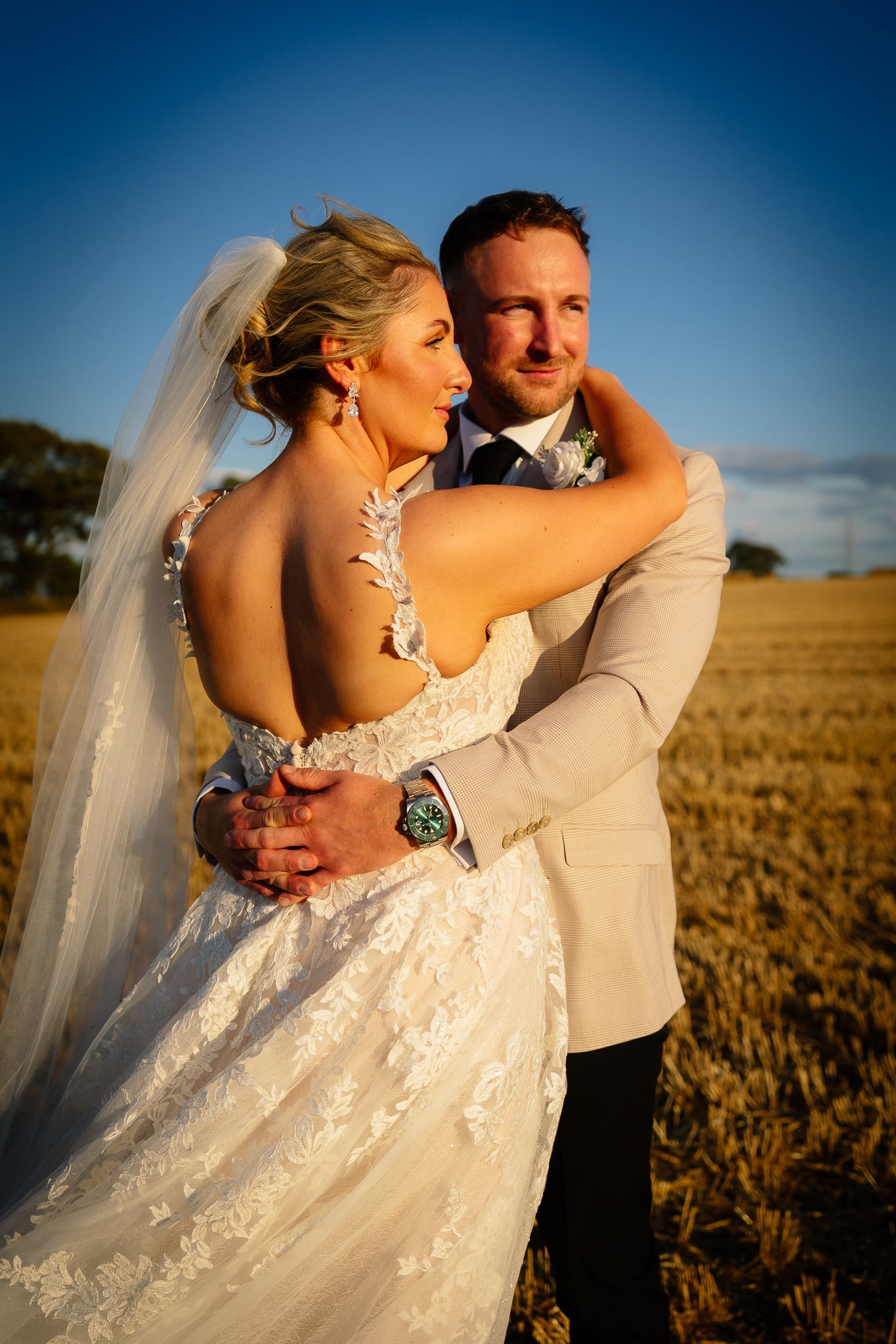 A romantic wedding couple embraces in a golden wheat field at sunset, with the bride's veil flowing in the breeze.