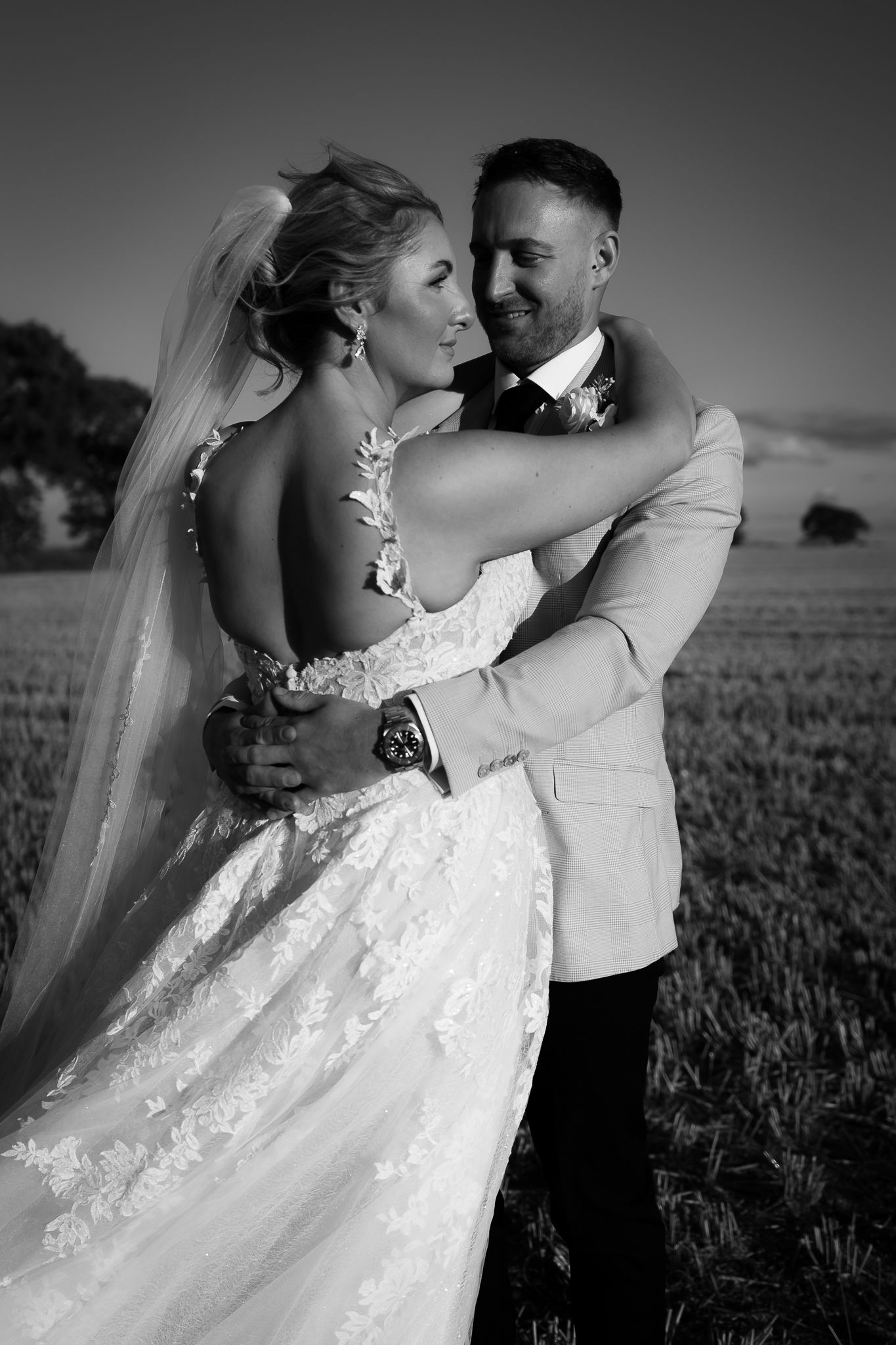 A black and white wedding portrait shows a couple in an intimate embrace among rural farmland.