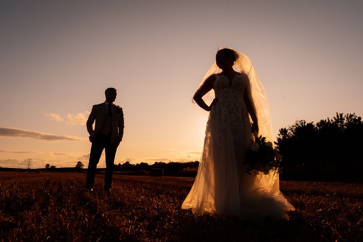 Silhouetted couple embraces at sunset in a golden field as their wedding dress flows in the evening breeze.