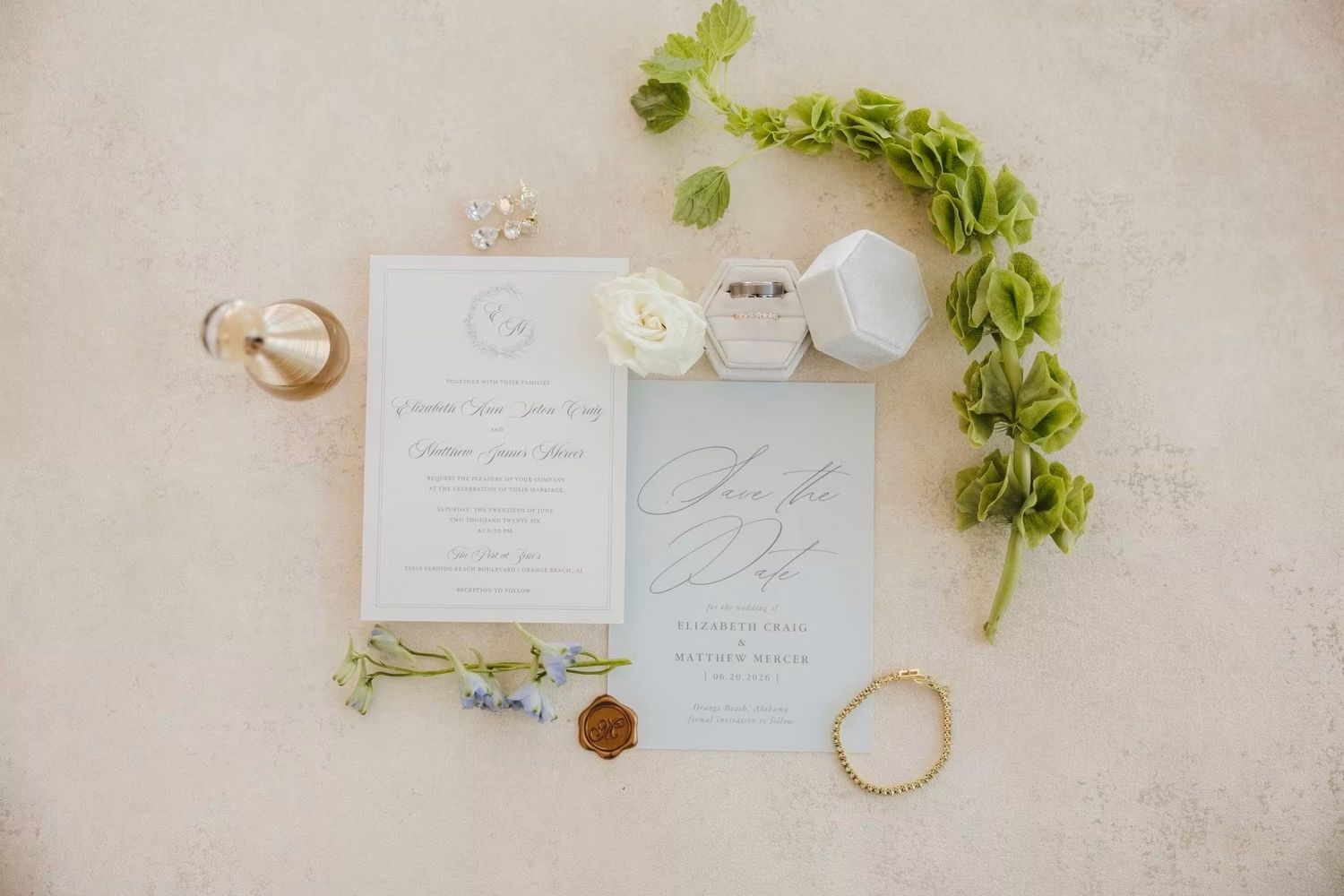 Elegant wedding stationery flat lay with blue invitation suite, gold rings, white florals, and greenery on neutral background.