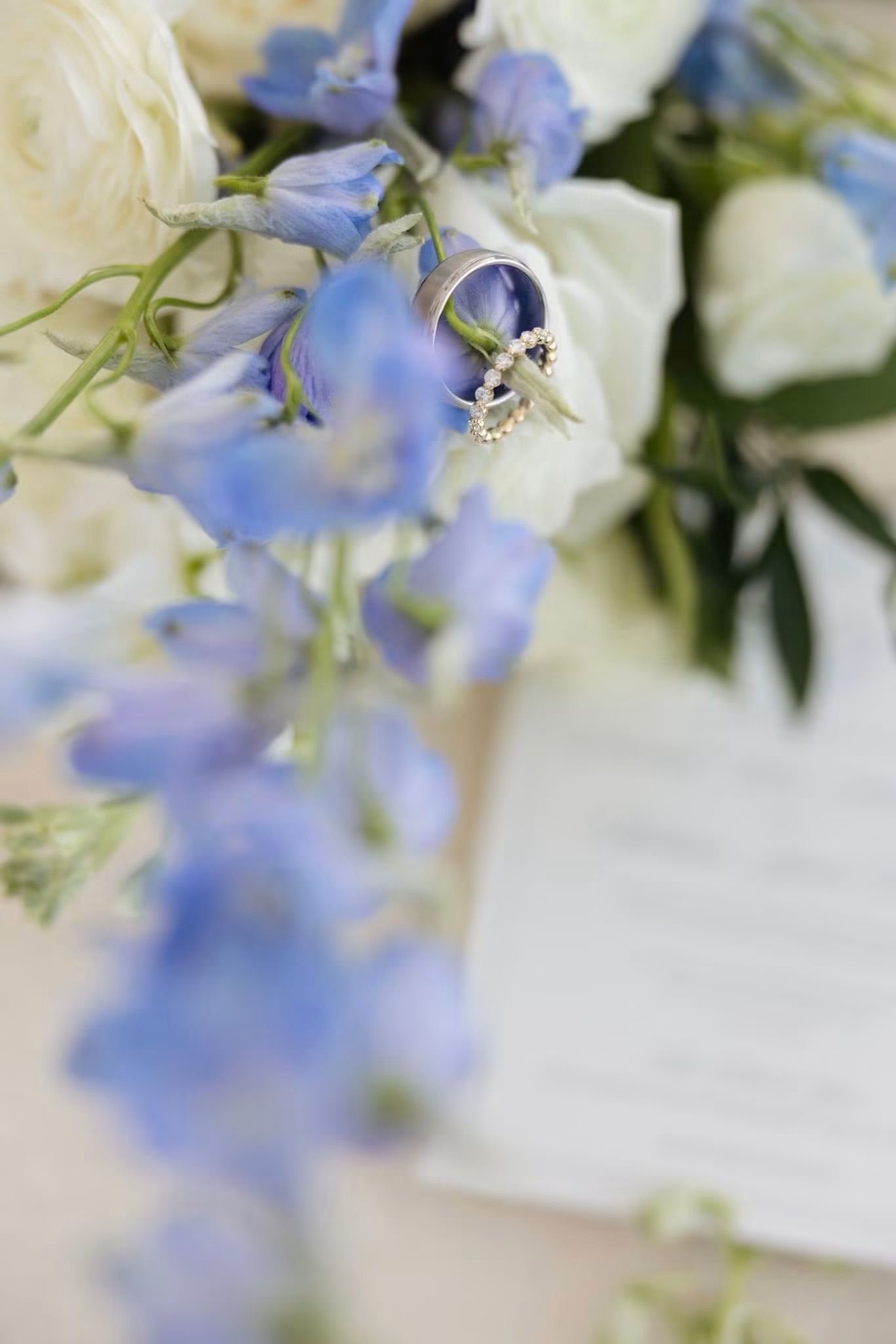 Wedding rings rest on blue delphinium and white roses bouquet with sheet music, elegant bridal details.